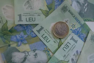Mai multe bancnote de un leu și o monedă sunt întinse pe o suprafață colorată cu motive florale.