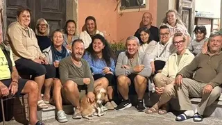 Un grup de persoane zâmbitoare stă împreună în fața unei clădiri, unele fiind așezate pe scaune, altele pe trepte.