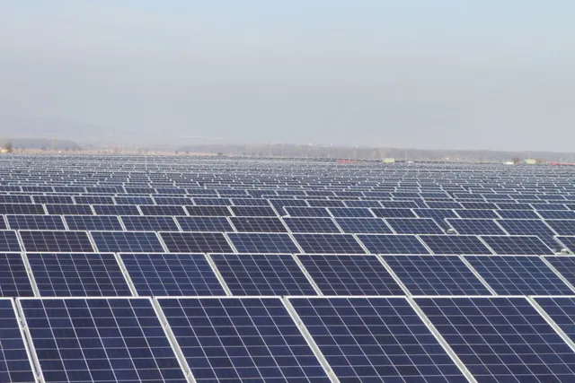 O întindere vastă de panouri solare fotovoltaice este dispusă pe un teren plat sub un cer înnorat.