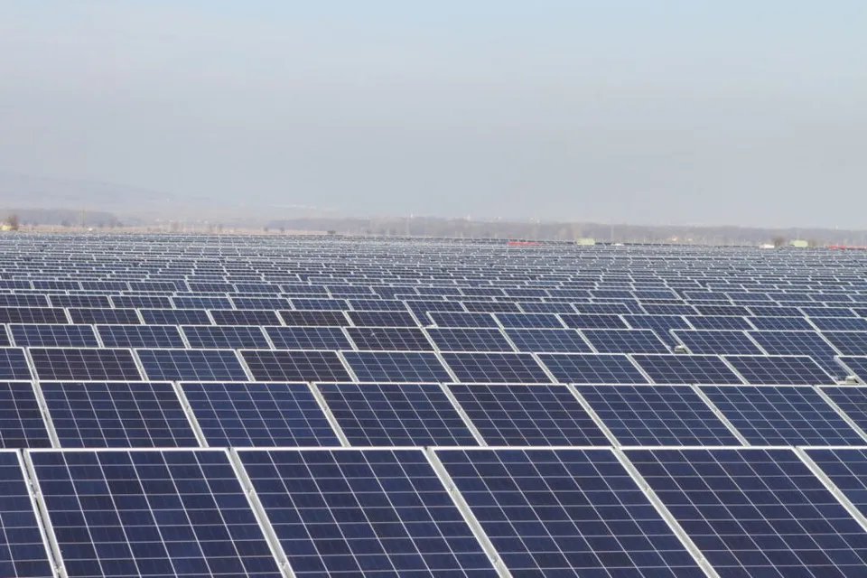 O întindere vastă de panouri solare fotovoltaice este dispusă pe un teren plat sub un cer înnorat.