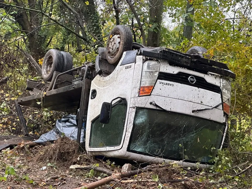Un camion alb este răsturnat pe o parte într-o zonă cu vegetație abundentă și copaci.