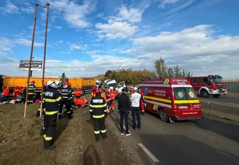 Inspectori în uniforme de protecție galben și negru, un vehicul de serviciu roșu și persoane pe marginea unui drum asfaltat sub un cer înnorat.