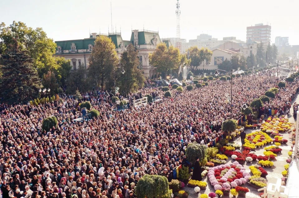O mulțime mare de oameni este adunată pe o stradă lată lângă clădiri și copaci, alături de straturi de flori colorate.