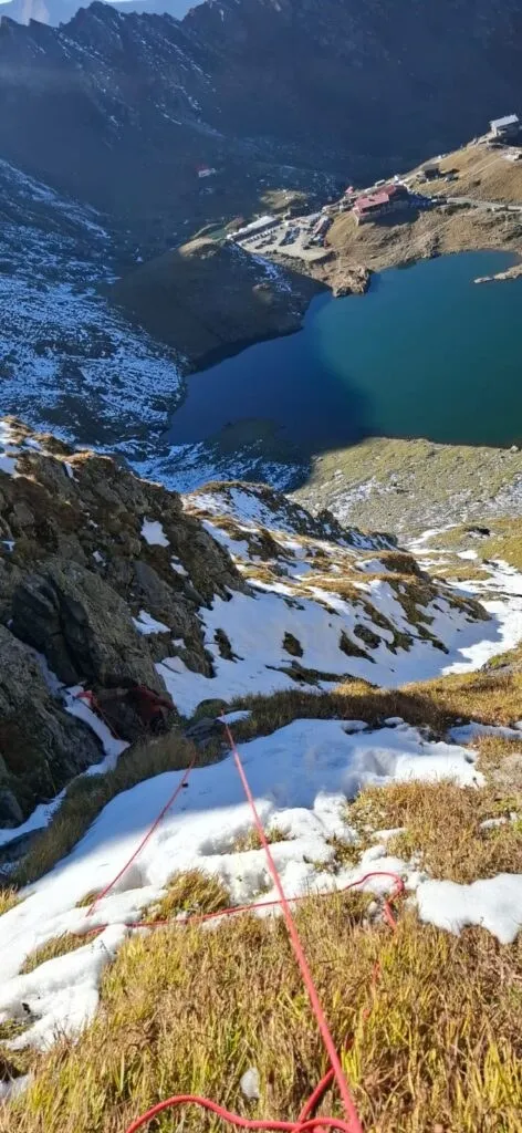 O coardă roșie întinsă pe un versant muntos acoperit de zăpadă și iarbă secă, cu un lac și munți în fundal.