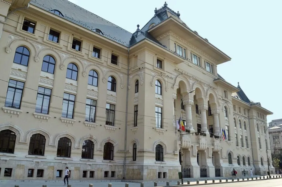 O clădire mare, de culoare deschisă, cu numeroase ferestre și elemente arhitecturale clasice, prezintă câteva steaguri suspendate pe fațadă.
