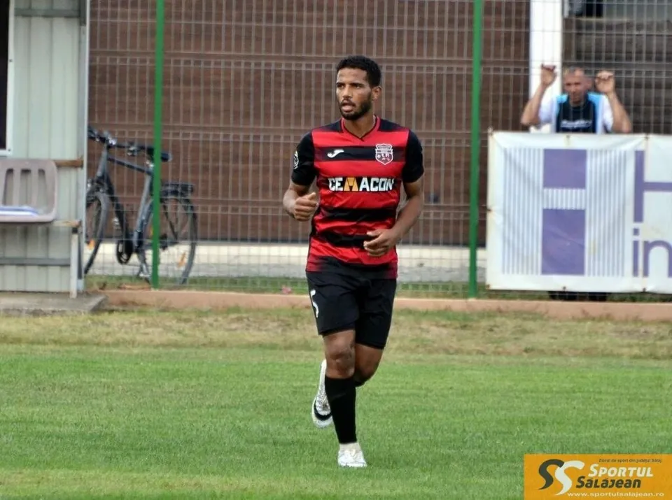 Un bărbat cu barbă poartă un tricou roșu și negru, pantaloni scurți negri și șosete negre, alergând pe un teren de fotbal cu iarbă verde.