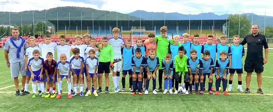 O echipă de juniori în echipamente sportive colorate stă în rând pe un teren de fotbal verde, alături de doi adulți.