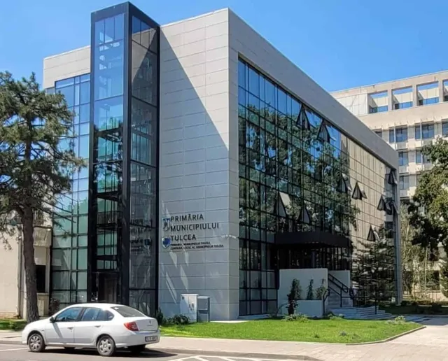 O clădire modernă cu fațadă de sticlă și panouri deschise este situată lângă un copac, având o mașină albă parcată în prim-plan.