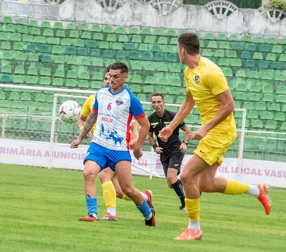 Câțiva jucători de fotbal în echipamente colorate aleargă pe un teren de gazon verde în fața unor tribune verzi.