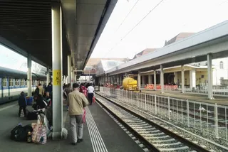 Oameni stau pe un peron de gară lângă bagaje, lângă șinele de tren și sub un acoperiș metalic de susținere.