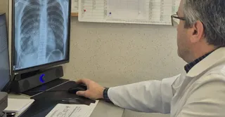 Un bărbat cu ochelari și halat alb stă la un birou utilizând un mouse în timp ce privește un monitor afișând o radiografie toracică.