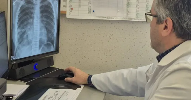 Un bărbat cu ochelari și halat alb stă la un birou utilizând un mouse în timp ce privește un monitor afișând o radiografie toracică.