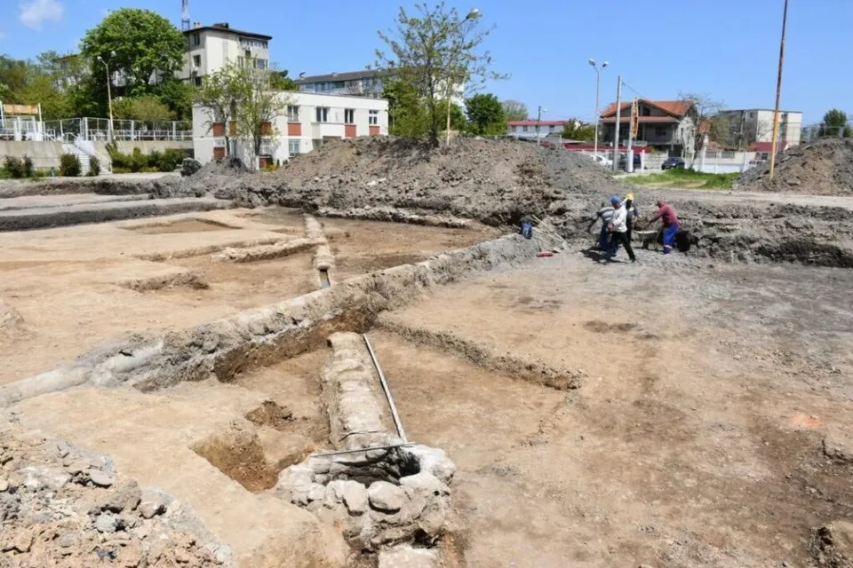 Oamenii lucrează într-un șantier de săpături arheologice unde se observă structuri de piatră și un tub subțire alungit în pământ.