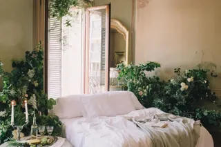 Un pat cu lenjerie albă este așezat într-o cameră decorată cu plante verzi și flori lângă o fereastră deschisă.