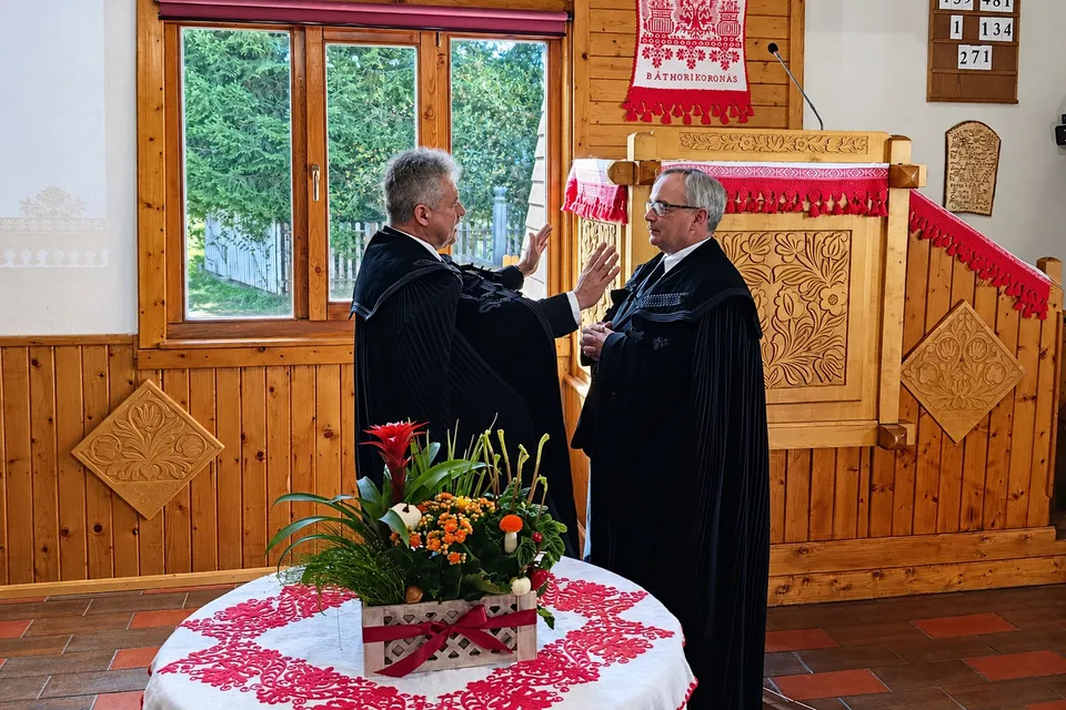 Doi bărbați îmbrăcați în robe negre stau în fața unui pupitru din lemn, lângă o masă rotundă cu un buchet de flori și un șervețel decorativ.