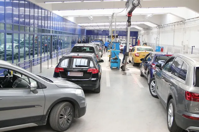 Mai multe mașini sunt parcate într-un spațiu interior cu podea albă și pereți albi, sub o lumină intensă.