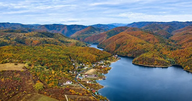 O vedere aeriană prezintă un lac înconjurat de dealuri acoperite de păduri cu frunziș colorat de toamnă și câteva clădiri mici la marginea apei.