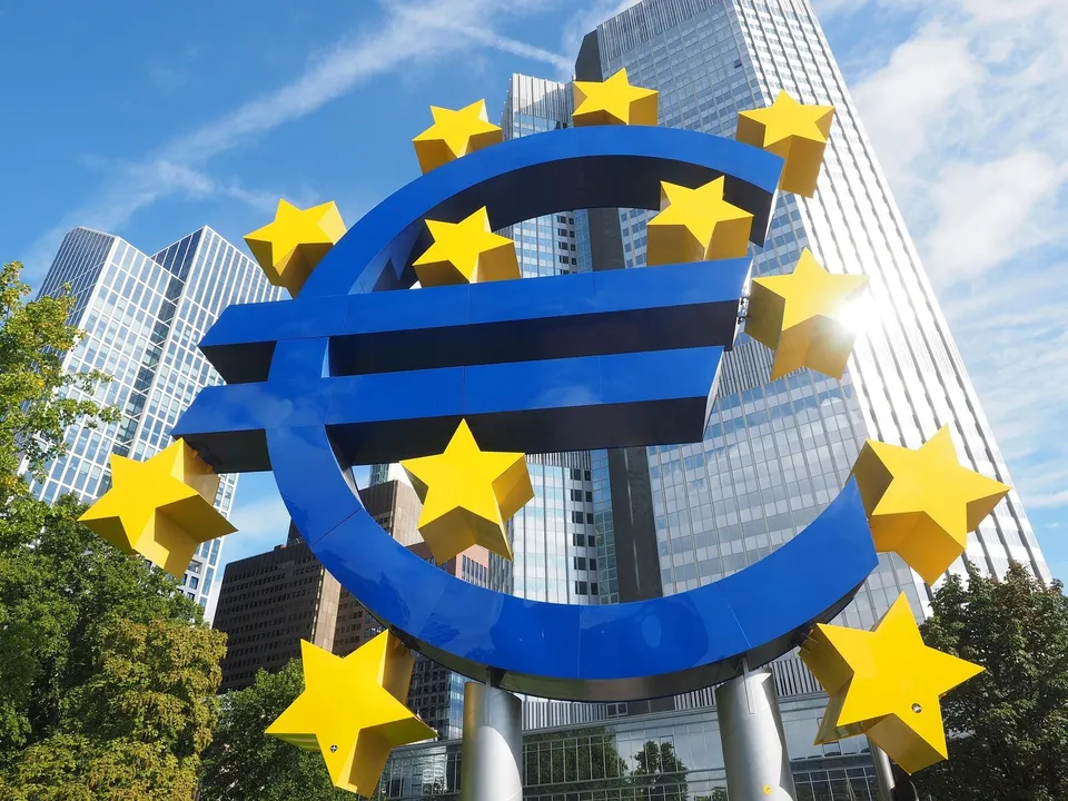 Un simbol Euro albastru cu stele galbene este amplasat în fața unor clădiri înalte de birouri sub un cer senin.