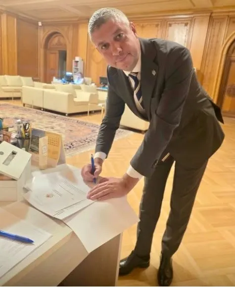 Un bărbat într-un costum gri și cravată stă aplecat peste un birou, ținând un pix deasupra unor hârtii albe într-o încăpere cu podea de lemn.