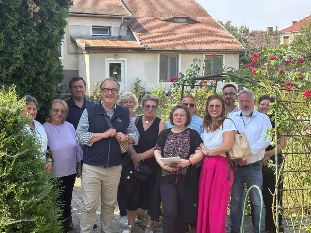 Un grup de persoane stă în picioare în fața unei clădiri cu acoperiș de țiglă, lângă un gard de plante și trandafiri.