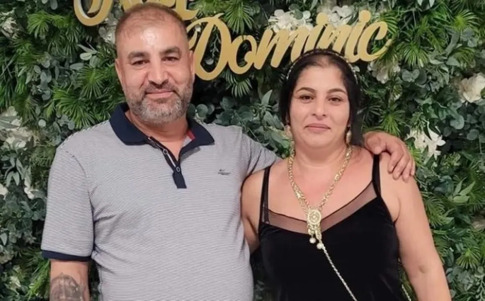 Un bărbat cu tricou polo gri și o femeie cu rochie neagră stau împreună în fața unui perete de plante verzi.