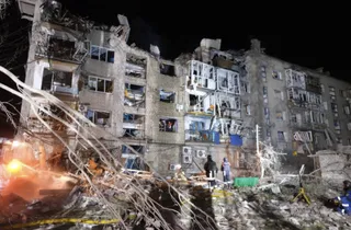 O clădire de apartamente cu ferestre distruse și fațadă deteriorată este iluminată noaptea lângă un morman de dărâmături și resturi.
