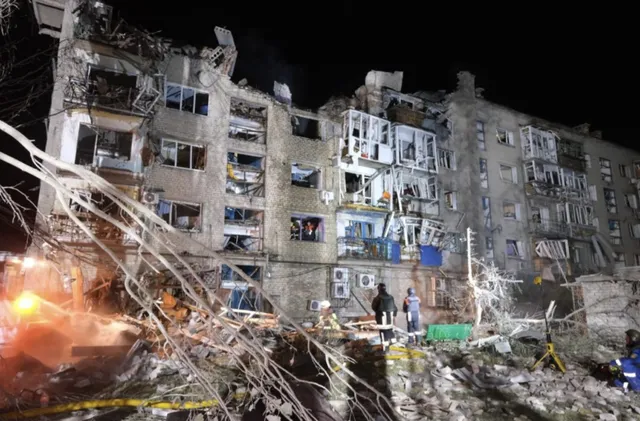 O clădire de apartamente cu ferestre distruse și fațadă deteriorată este iluminată noaptea lângă un morman de dărâmături și resturi.