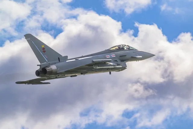 Un avion de luptă gri zboară pe un cer albastru cu nori albi.