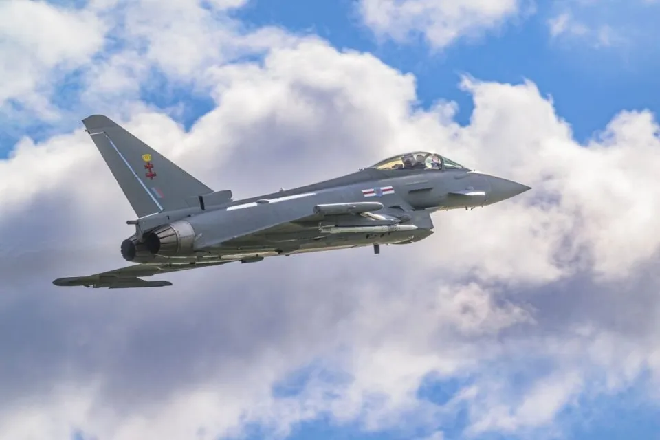 Un avion de luptă gri zboară pe un cer albastru cu nori albi.
