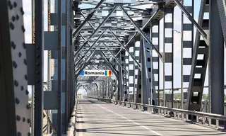 O structură metalică de pod cu grinzi și stâlpi gri formează un tunel peste o suprafață de rulare asfaltată cu linii albe.