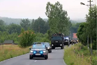 Un convoi format din vehicule militare și o mașină de poliție circulă pe un drum asfaltat înconjurat de vegetație și copaci.