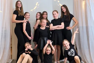 Opt fete îmbrăcate în haine negre poză în grup în fața unor draperii albe.