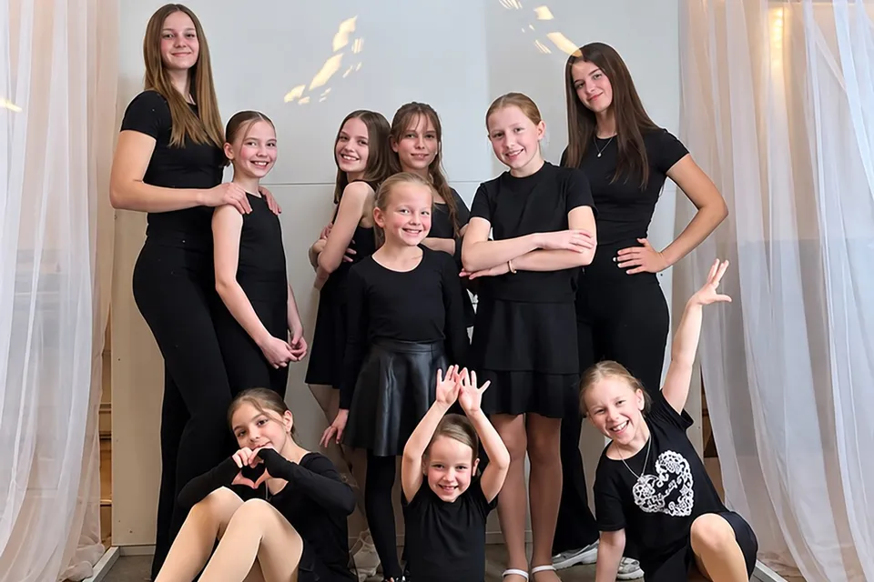 Opt fete îmbrăcate în haine negre poză în grup în fața unor draperii albe.