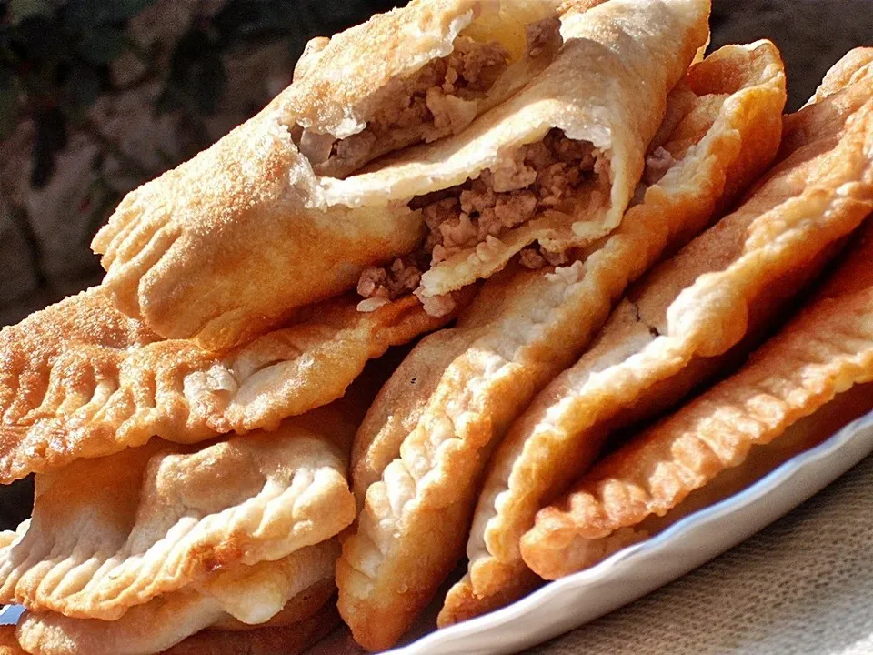 Mai multe produse de patiserie prăjite, cu margini zimțate și umplutură de carne, sunt așezate pe o farfurie albă.
