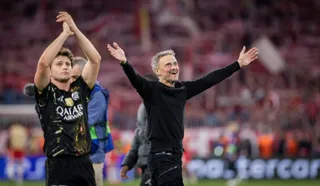Un bărbat cu părul cărunt și haine negre stă cu brațele întinse în lateral pe un teren de fotbal, în timp ce un jucător aplaudă în fundal.