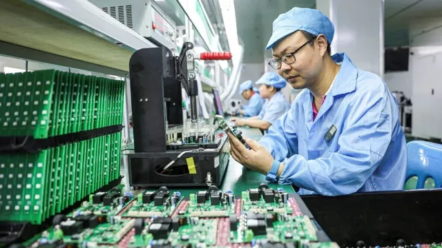 Un bărbat cu ochelari și căciulă de protecție albastră examinează componente electronice pe o masă de lucru într-o linie de producție.