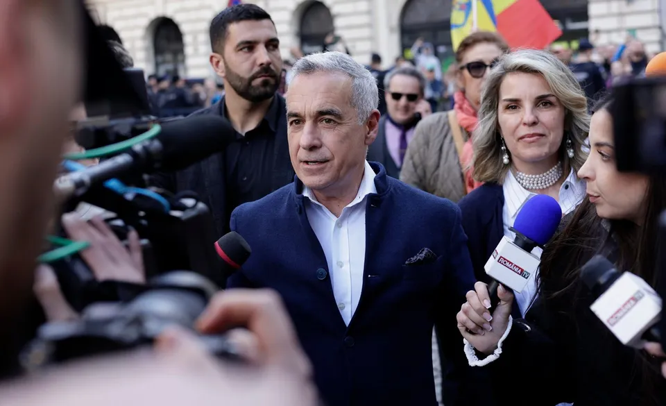 Un bărbat cu părul cărunt și sacou albastru vorbește în fața mai multor microfoane și camere de filmare într-un grup de oameni.