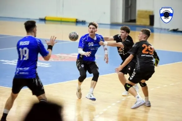 Un jucător de handbal în echipament albastru sare pe teren, încercând să arunce mingea spre adversari în timpul unui meci de sală.
