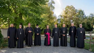 Zece bărbați îmbrăcați în robe negre și unele roba cu bandă roz stau în rând pe o cărare pavată în fața unor copaci verzi.