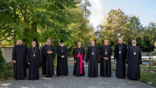 Zece bărbați îmbrăcați în robe negre și unele roba cu bandă roz stau în rând pe o cărare pavată în fața unor copaci verzi.