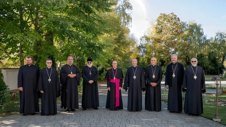Zece bărbați îmbrăcați în robe negre și unele roba cu bandă roz stau în rând pe o cărare pavată în fața unor copaci verzi.