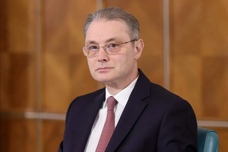 Un bărbat cu ochelari, purtând sacou închis la culoare, cămașă albă și cravată roz, stă în fața unui fundal din panouri de lemn maro.