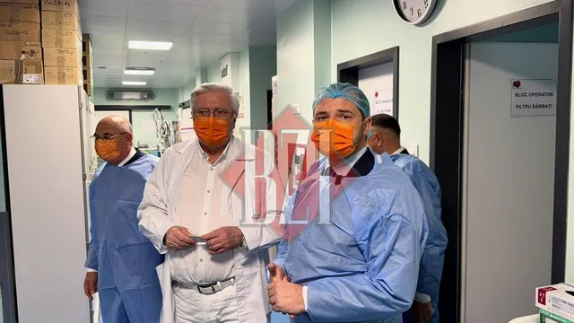 Patru bărbați purtând măști și halate medicale stau într-un hol de spital lângă un ușă și un ceas de perete.