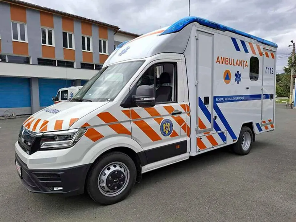 O ambulanță albă cu dungi portocalii și albastre este parcată pe un asfalt gri în fața unei clădiri cu ferestre albe.