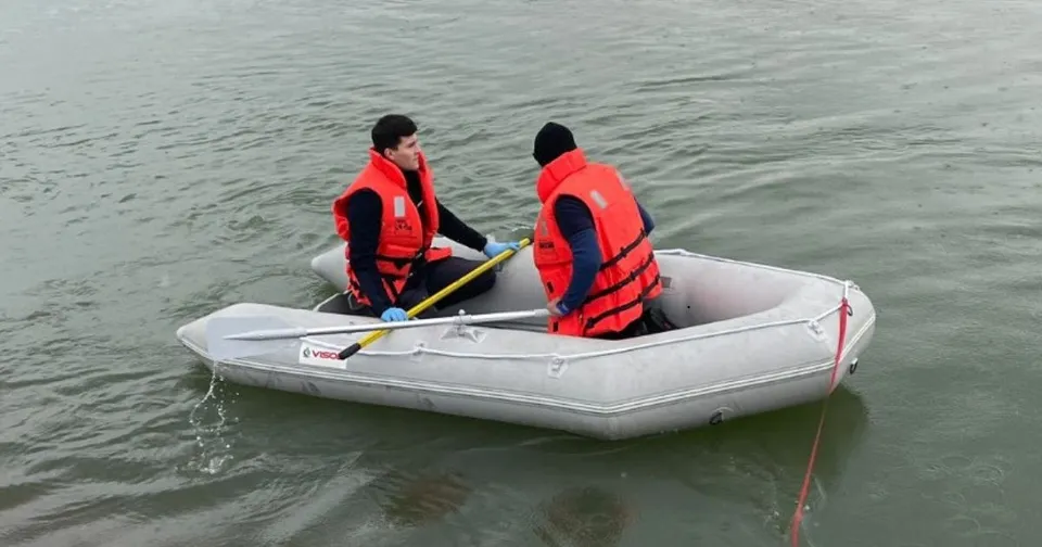 Două persoane purtând veste salvatoare portocalii se află într-o barcă gonflabilă gri pe un plan de apă.
