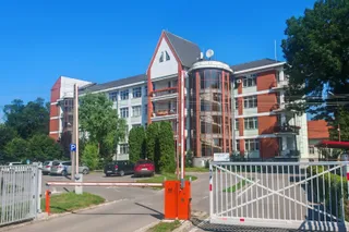 O clădire mare cu fațadă albă și elemente roșii este situată în spatele unui gard metalic și al unei bariere de acces.