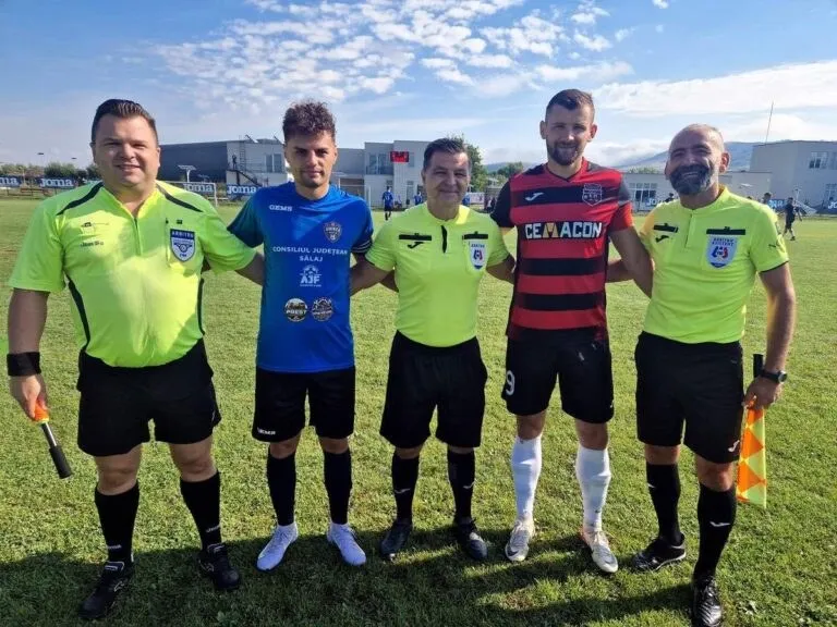 Cinci bărbați stau în rând pe un teren de fotbal, purtând tricouri colorate și șorturi scurte, sub un cer cu nori dispersați.