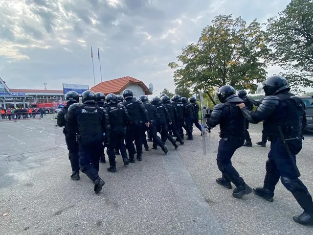 O grupă de persoane purtând uniforme negre și echipamente de protecție se deplasează pe un drum asfaltat lângă un grup de copaci.