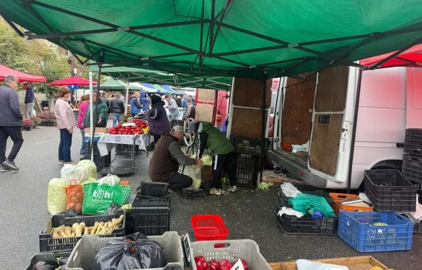 O piață acoperită cu un cort verde, cu oameni care vând și cumpără produse, cu diverse cutii, cosuri și saci pline de marfă.