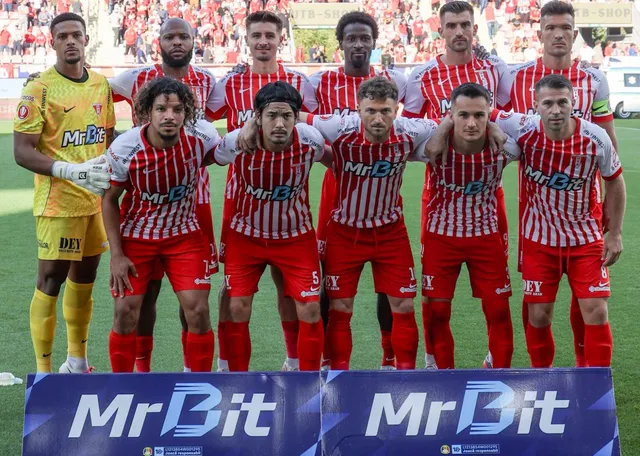 O echipă de fotbaliști în echipamente roșii și albe stă în rând, în timp ce un jucător în echipament galben stă în lateral.